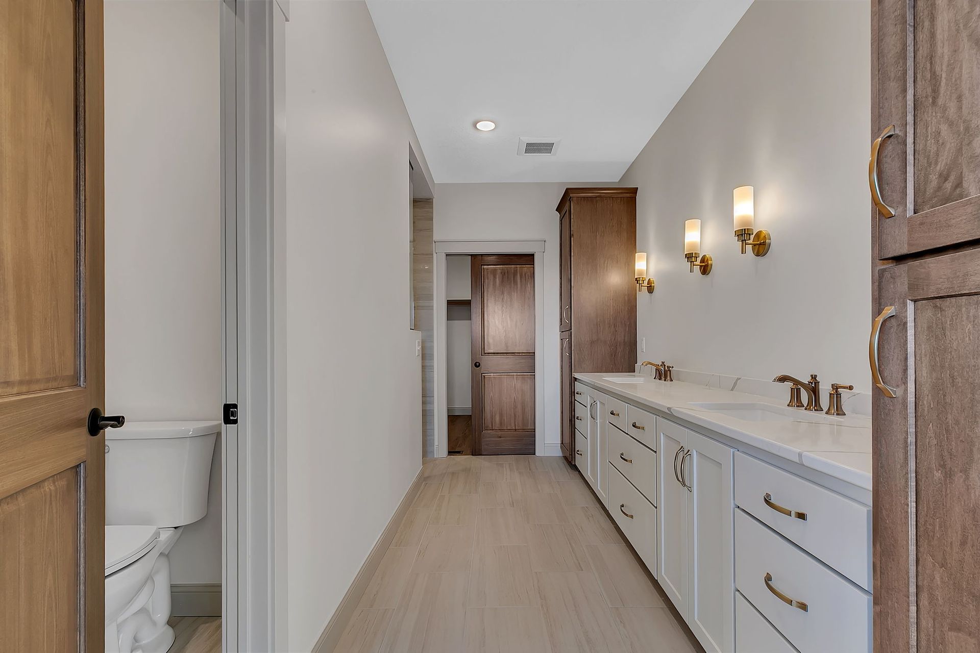 Bathroom with white walls, long counter with sinks, wooden cabinets, and toilet.