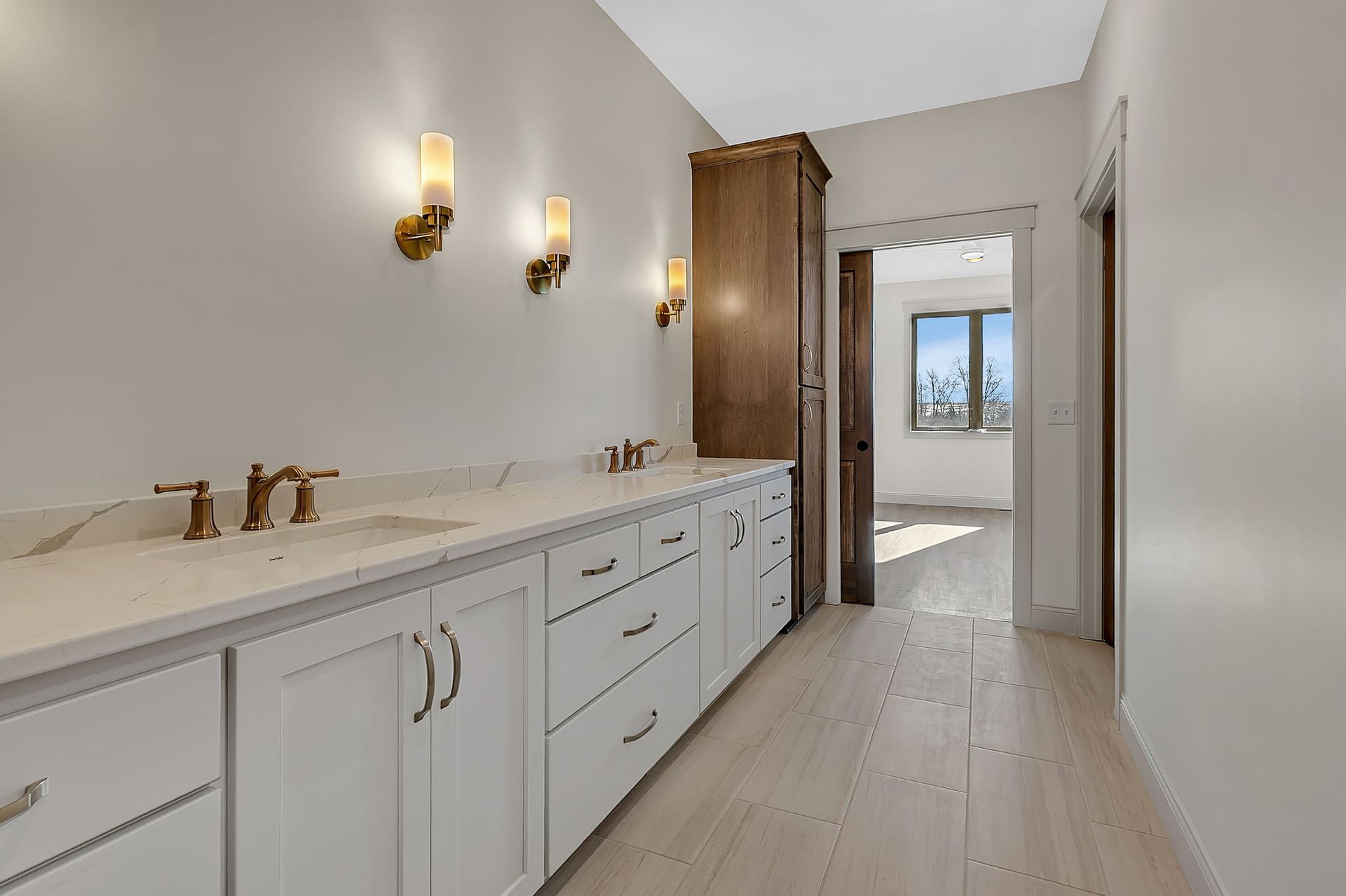 A bathroom with a white vanity, gold fixtures, and sconces. Doorway leads to a bedroom.