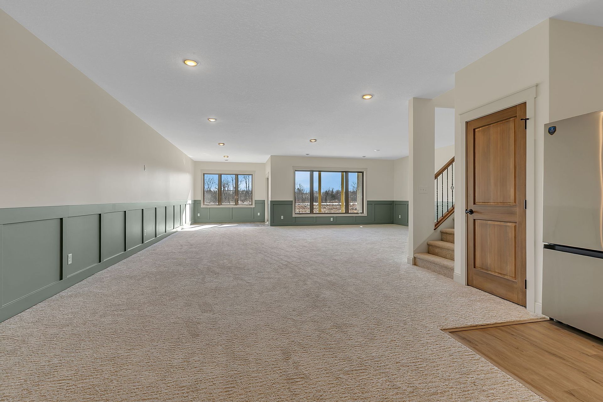 Empty carpeted room with light green wainscoting, windows, staircase, and a wooden door.
