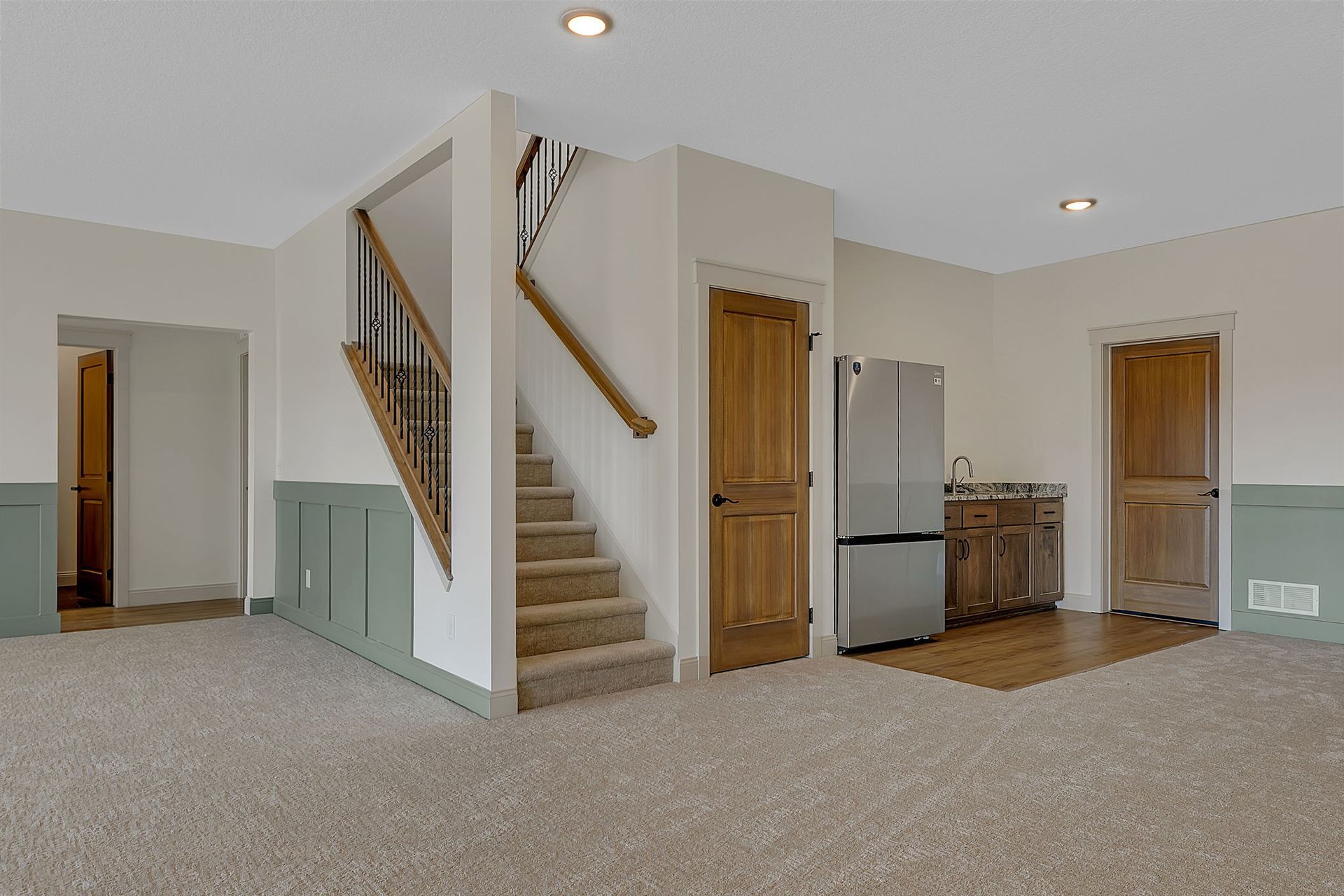 Beige carpeted room with stairs, two wooden doors, a mini-fridge, and green paneling.