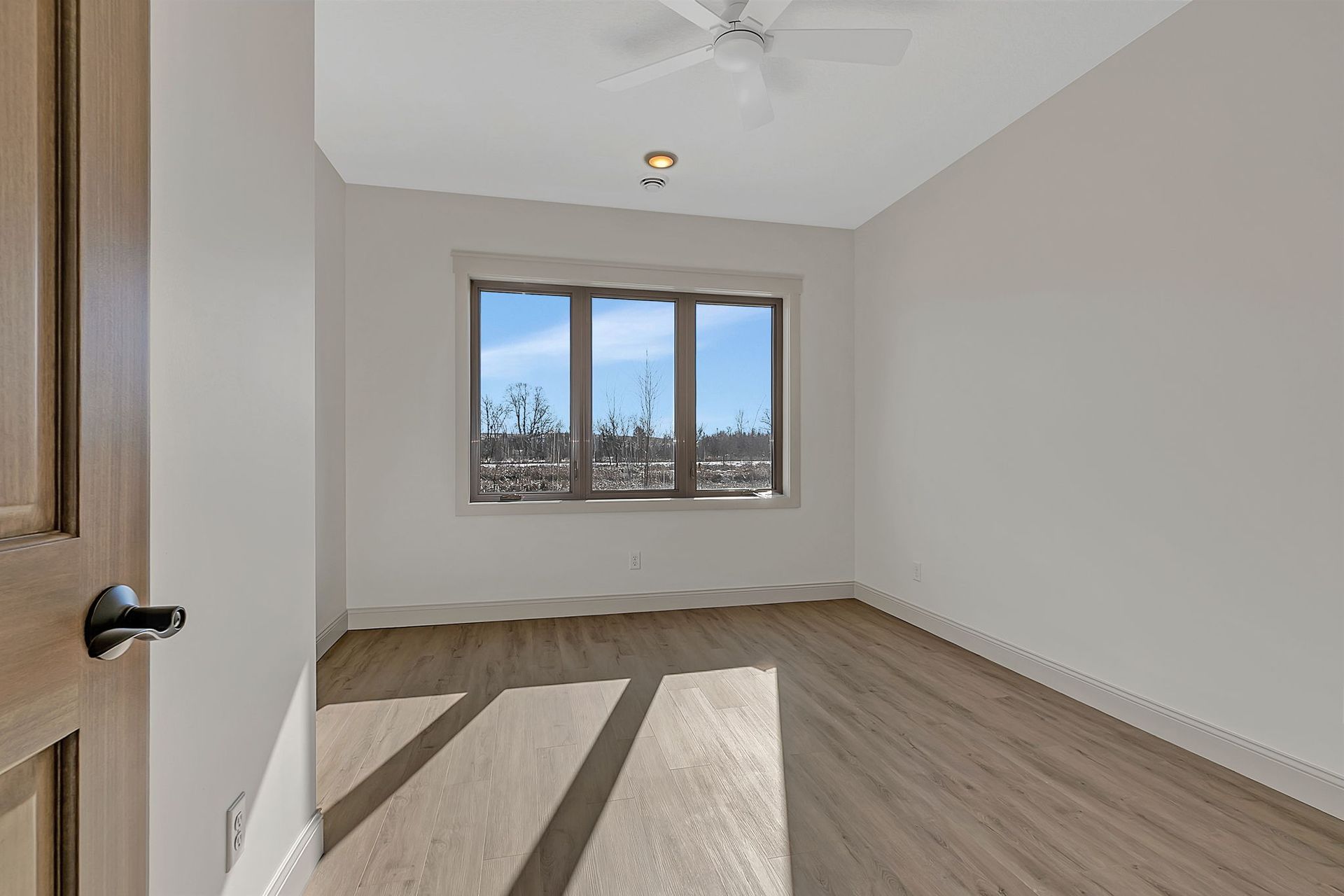 Empty room with window, wood floors, and neutral walls. Door on left.
