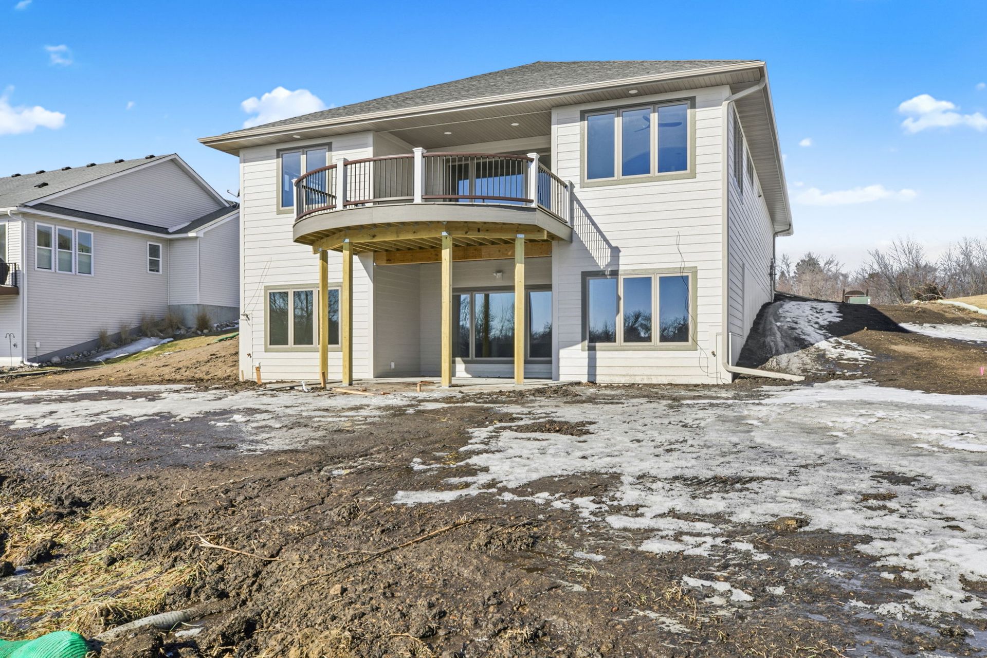 Two-story house with balcony and ground-level patio, set in a muddy yard under a cloudy sky.