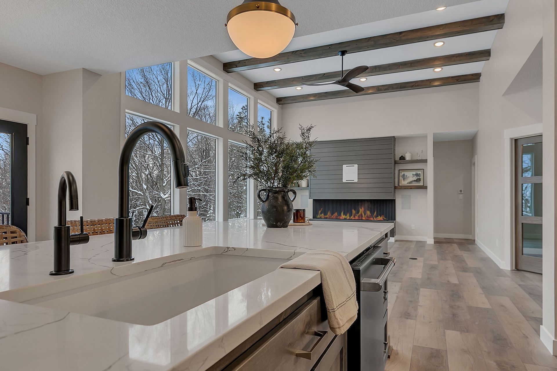Modern kitchen with white countertops, large windows overlooking snowy trees, and a fireplace.