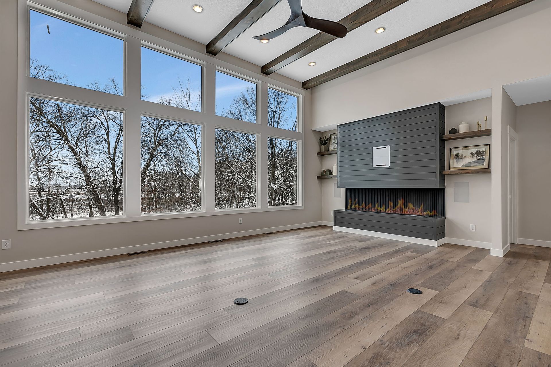 Spacious room with large windows, fireplace, wood beams, and hardwood floors; view of trees outside.