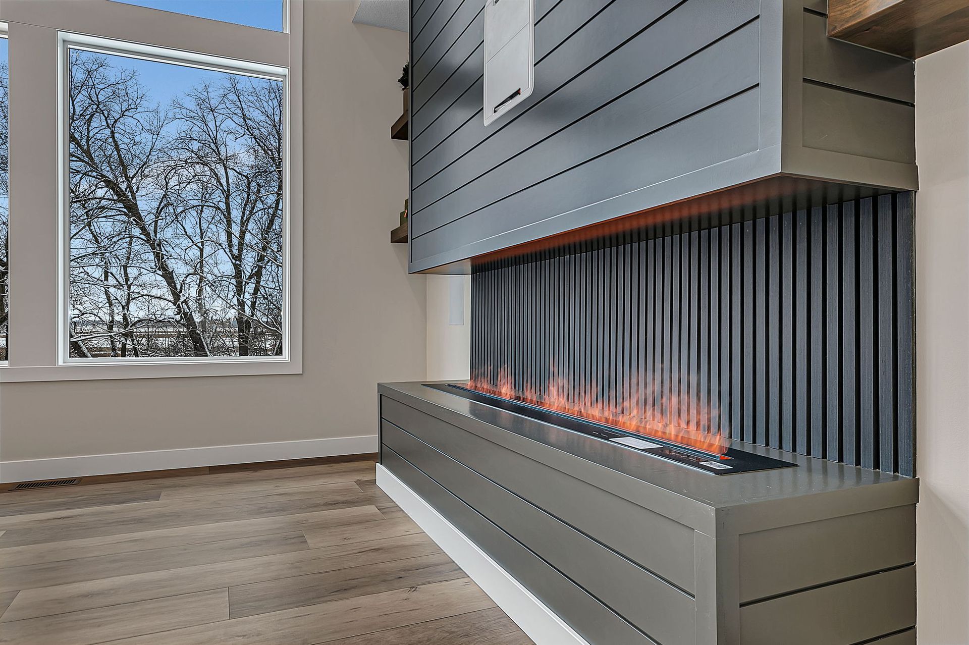 Modern fireplace with gray paneling and vertical flame effect, next to a window with a view of bare trees.