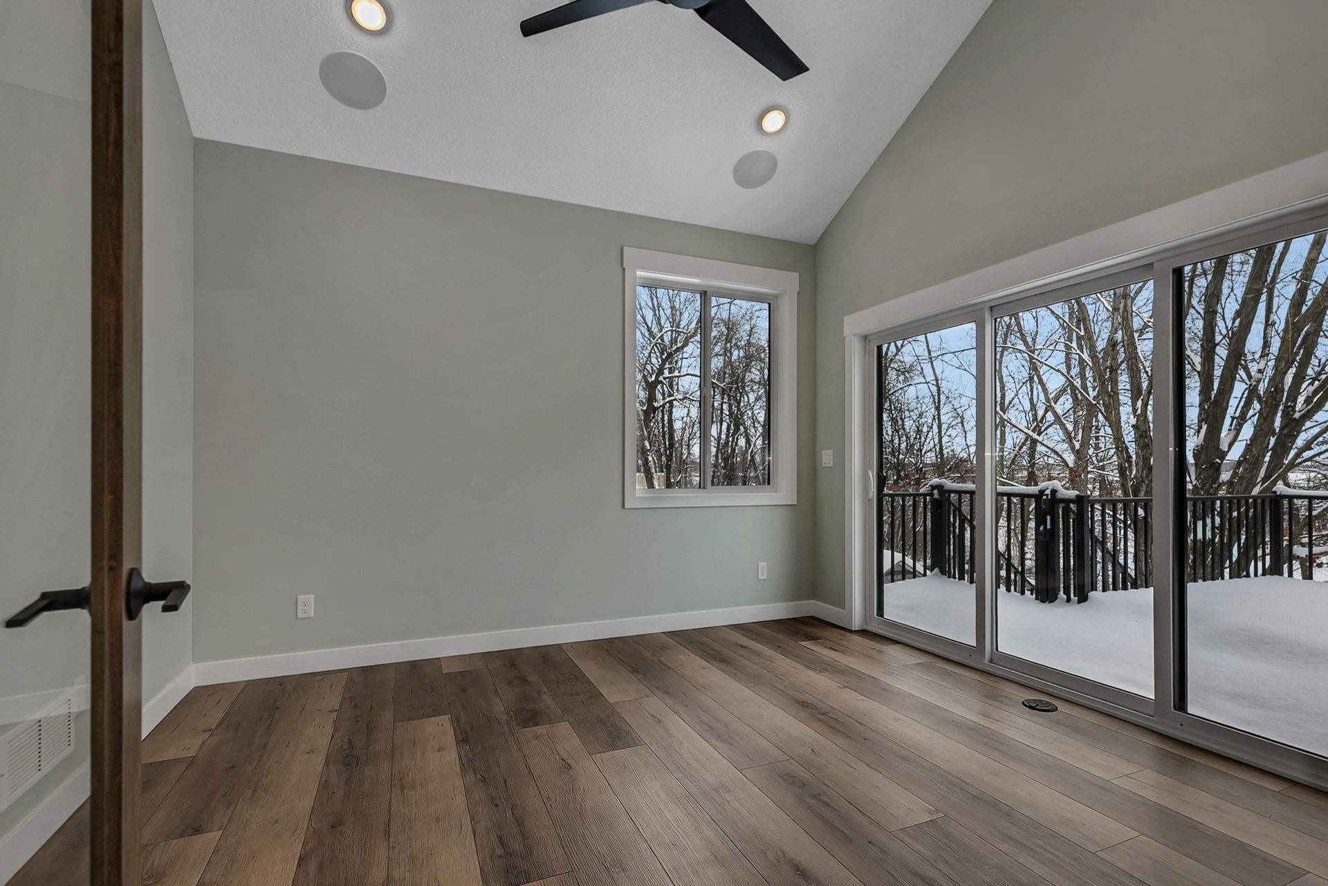 Bedroom with wooden floor, sliding glass door to deck, window, light gray walls, and vaulted ceiling.