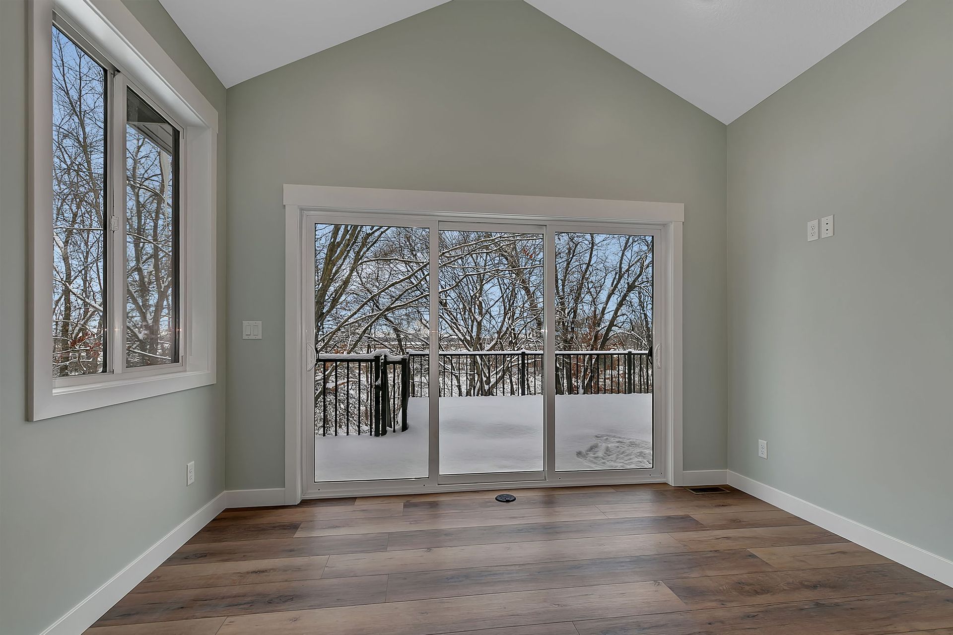 Empty room with large sliding glass door and window, overlooking a snowy deck and trees.
