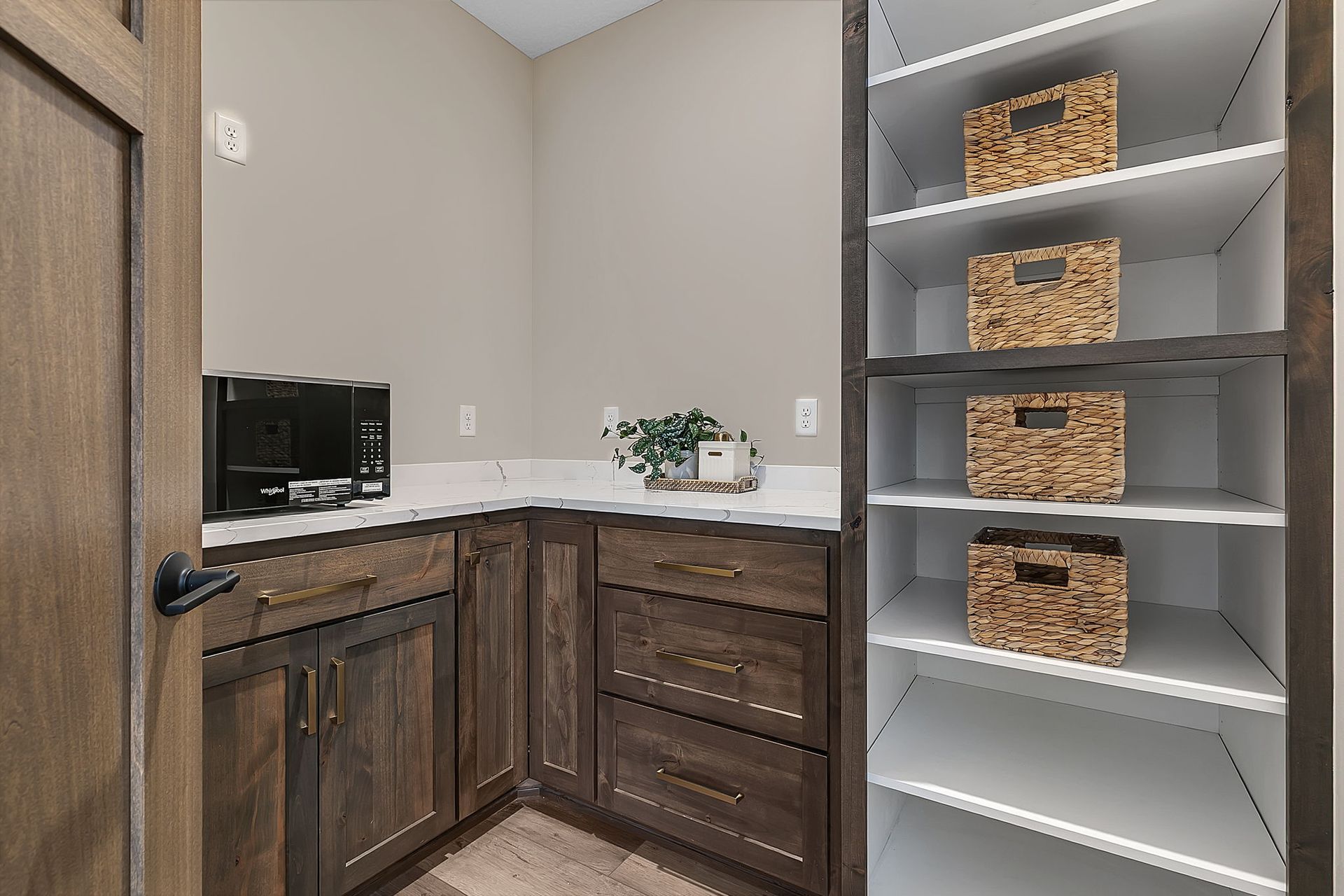 Pantry with dark wood cabinets, white countertop, shelving with woven baskets, and a coffee maker.