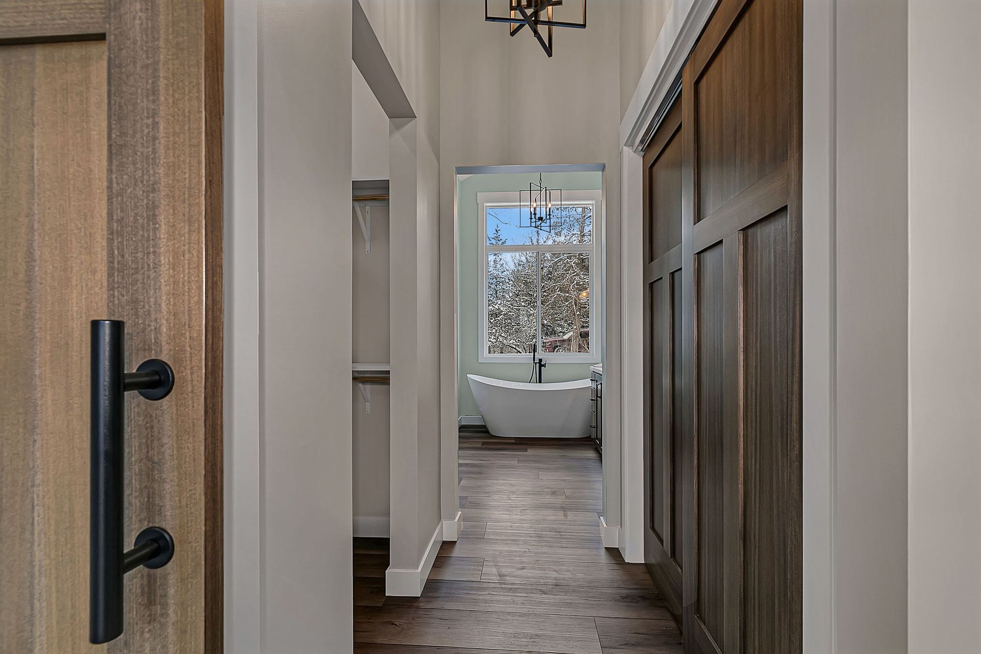 Hallway with wooden door, dark flooring, leading to a bathroom with a window and a bathtub.