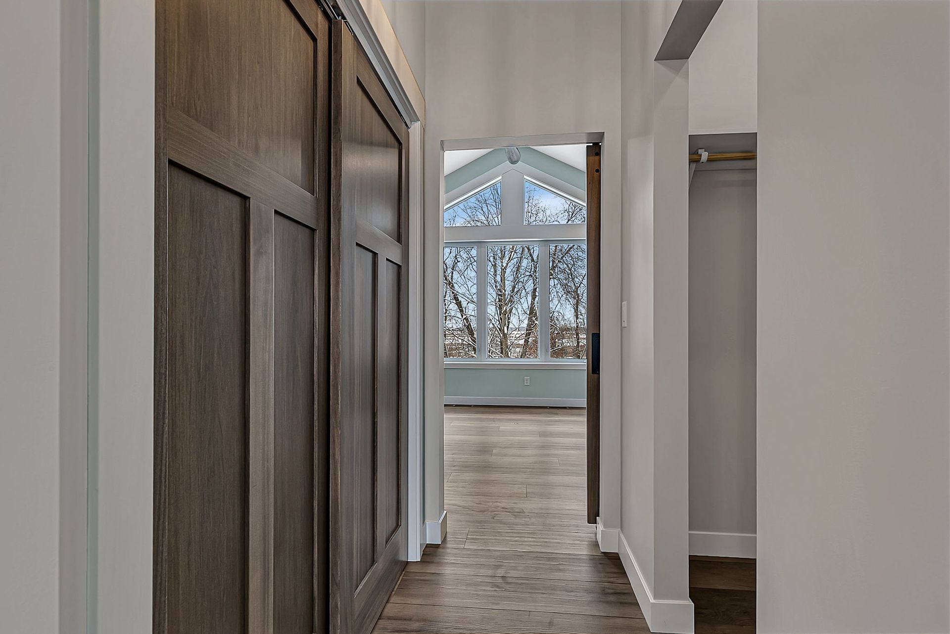 Hallway with dark wood closet doors and a room with large windows at the end.