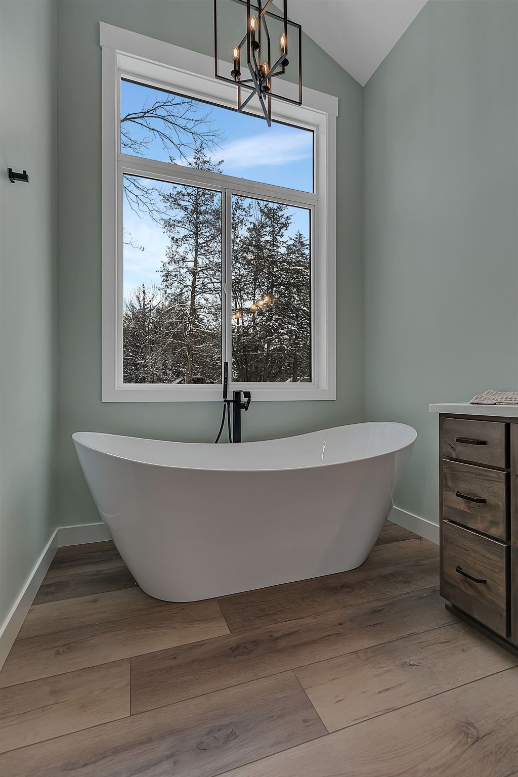 Freestanding white bathtub under large window in a light green bathroom with wood-look flooring.