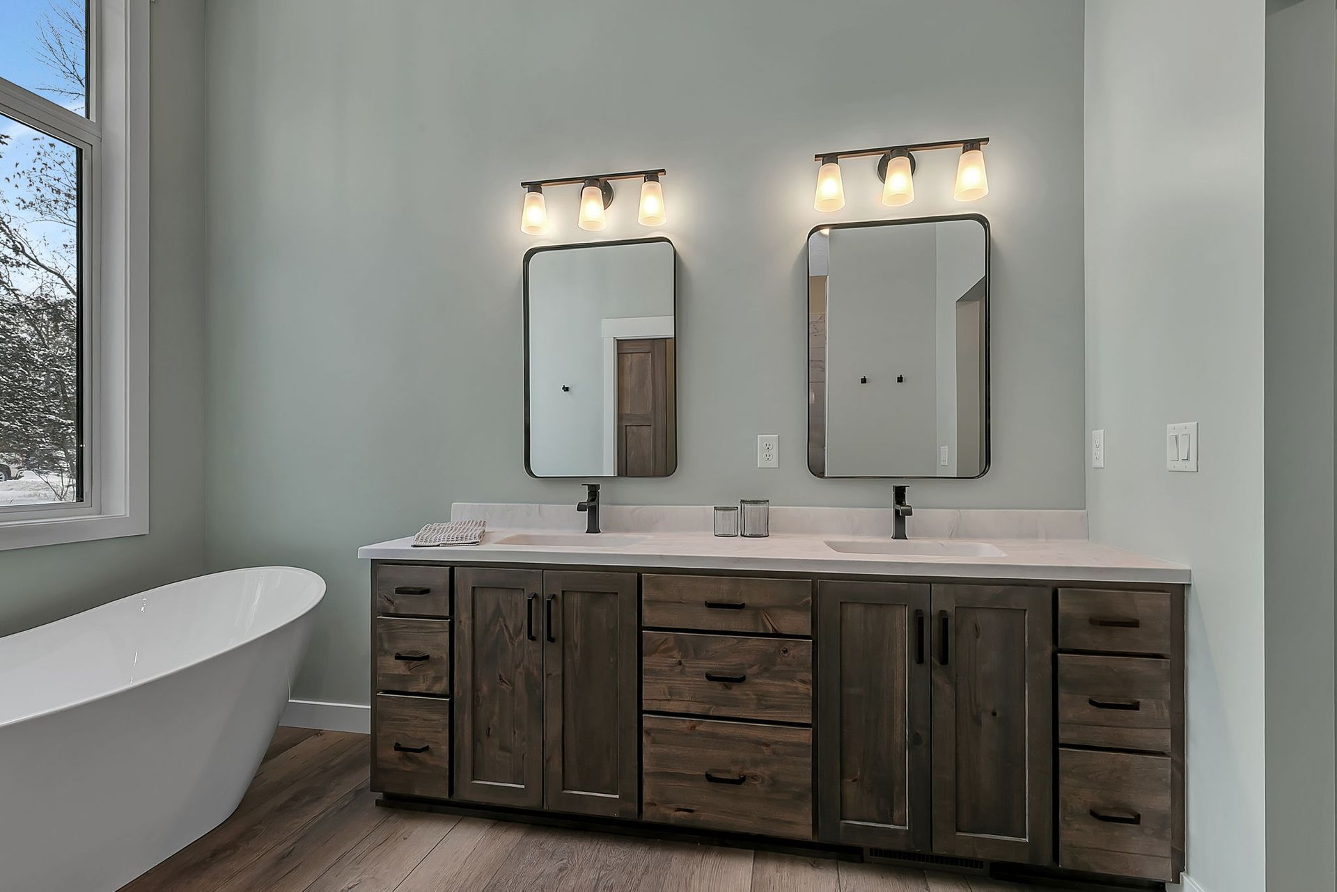 Bathroom with double vanity, two mirrors, and a freestanding tub. Gray walls, wooden cabinets, and light-colored countertops.