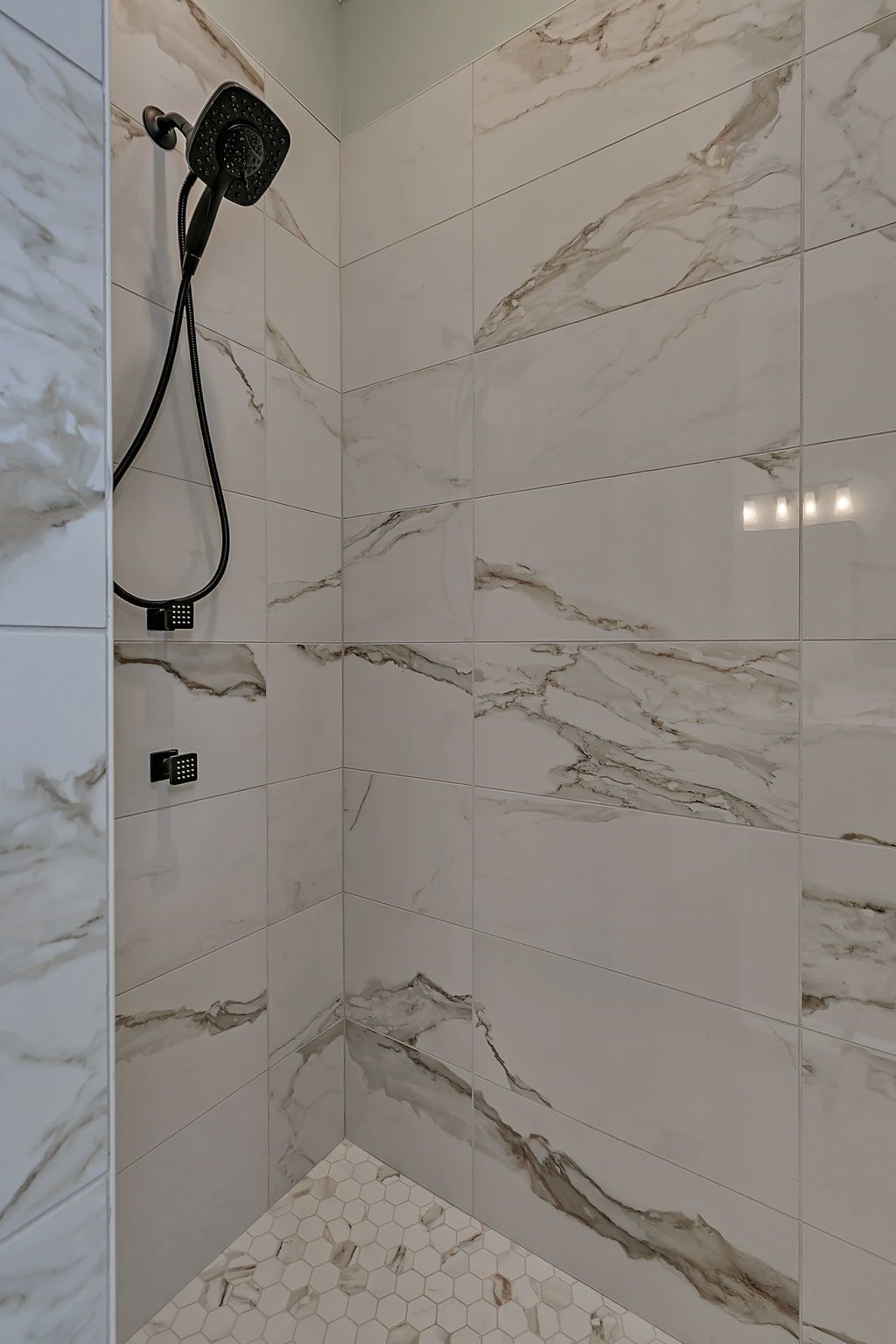 Shower stall with white marble-look tiles and a black showerhead. The floor has small, round, white tiles.