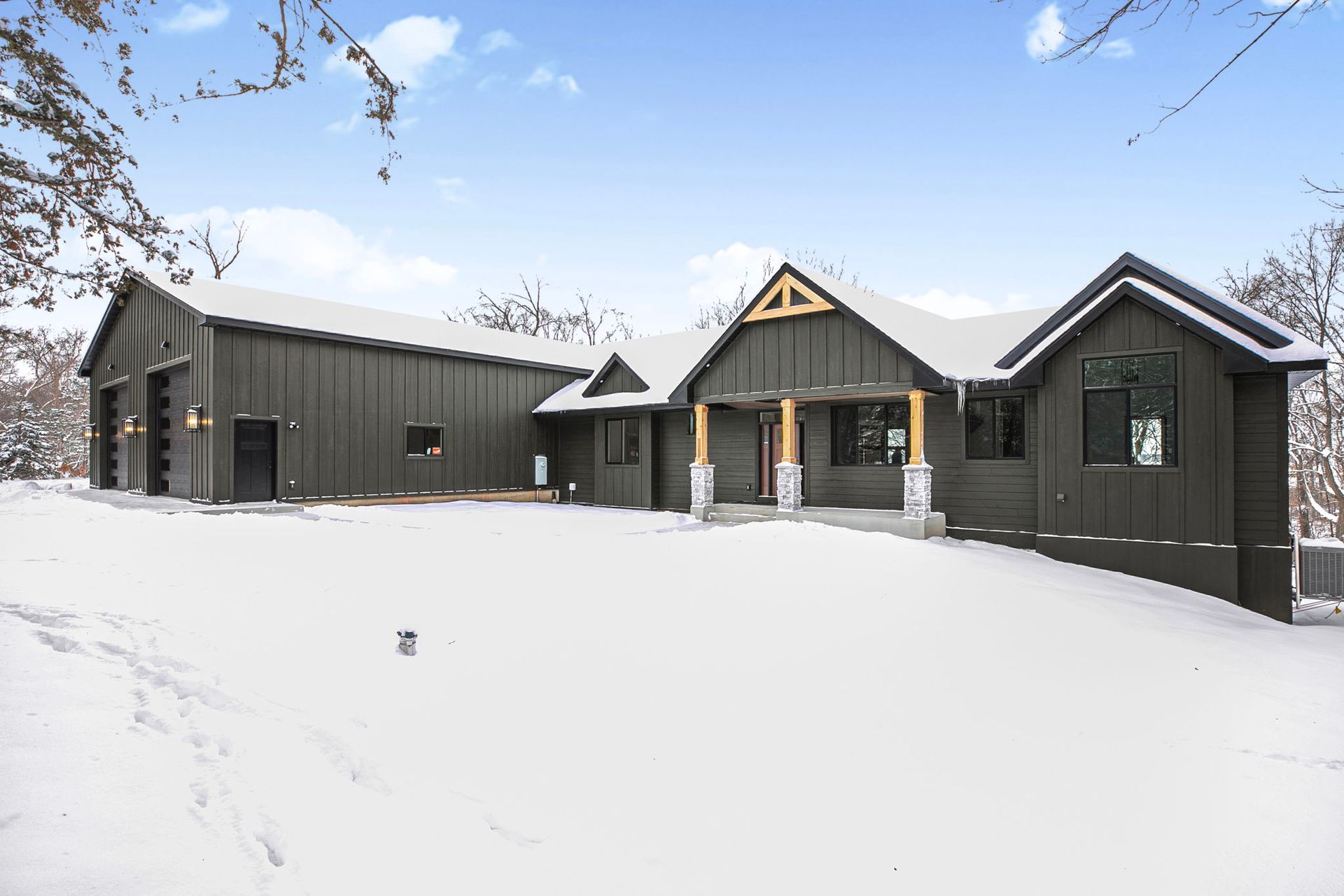 Dark gray house and garage in a snowy landscape. Wooden trim, large windows, overcast sky.