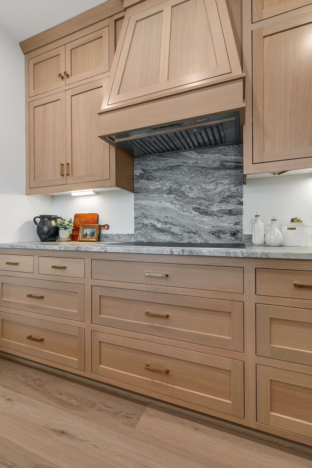Light wood kitchen cabinets, marble countertop, and a gray stone backsplash with a central range hood.