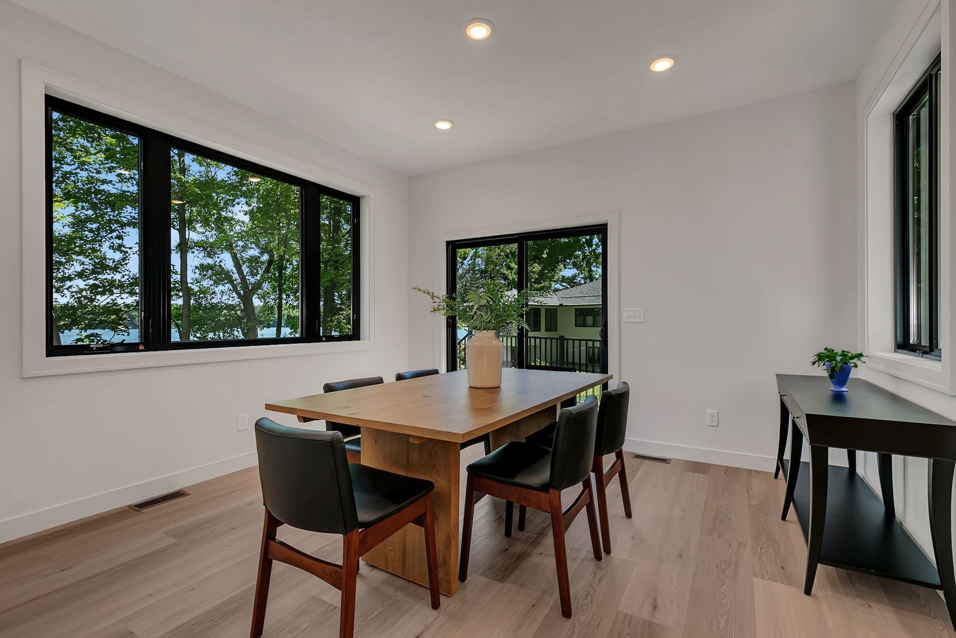 Bright dining room with wooden table, black chairs, large windows, and a side desk with a plant.
