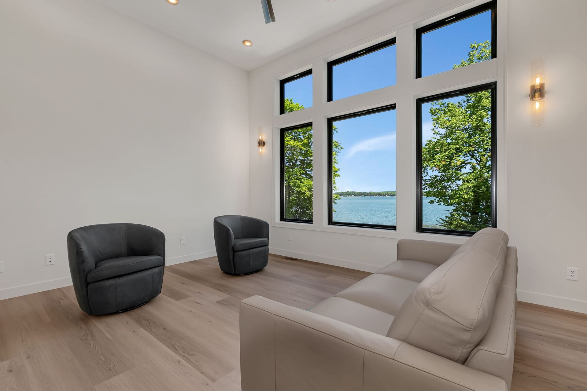 Bright modern living room with large windows, light wood floors, and two dark chairs facing a beige sofa.