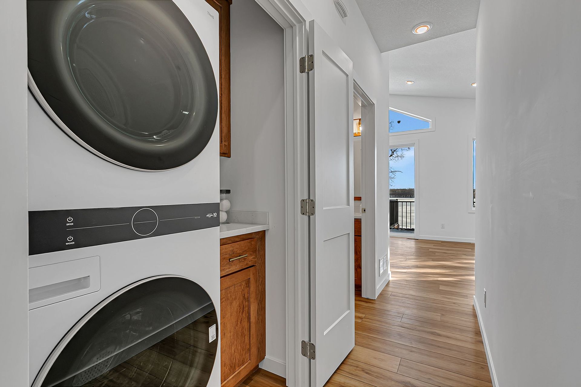Stacked washer and dryer in a laundry room with hardwood floors and a hallway leading to another room.