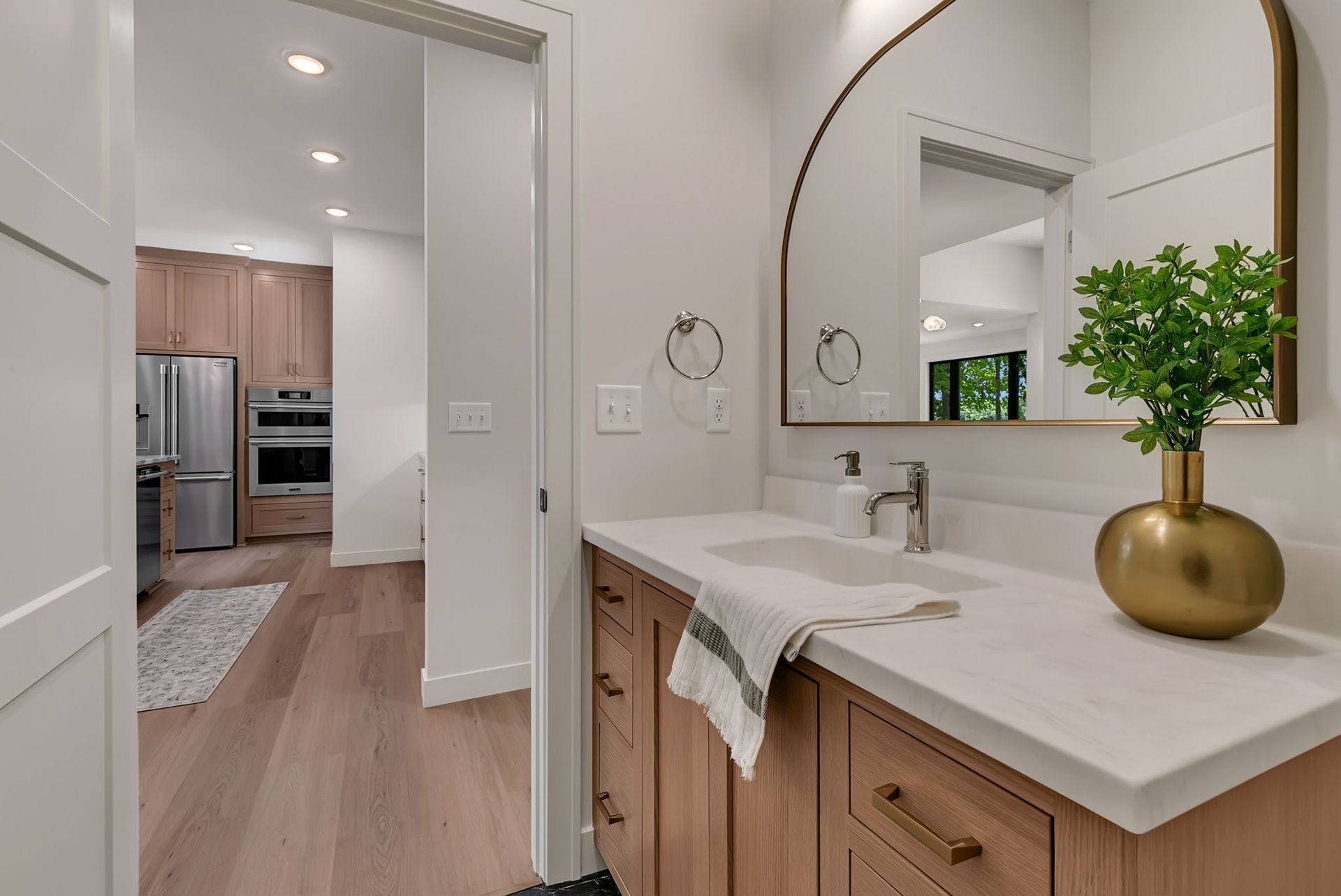 Bright bathroom vanity with arched mirror, wood cabinets, and hallway view to a kitchen beyond.