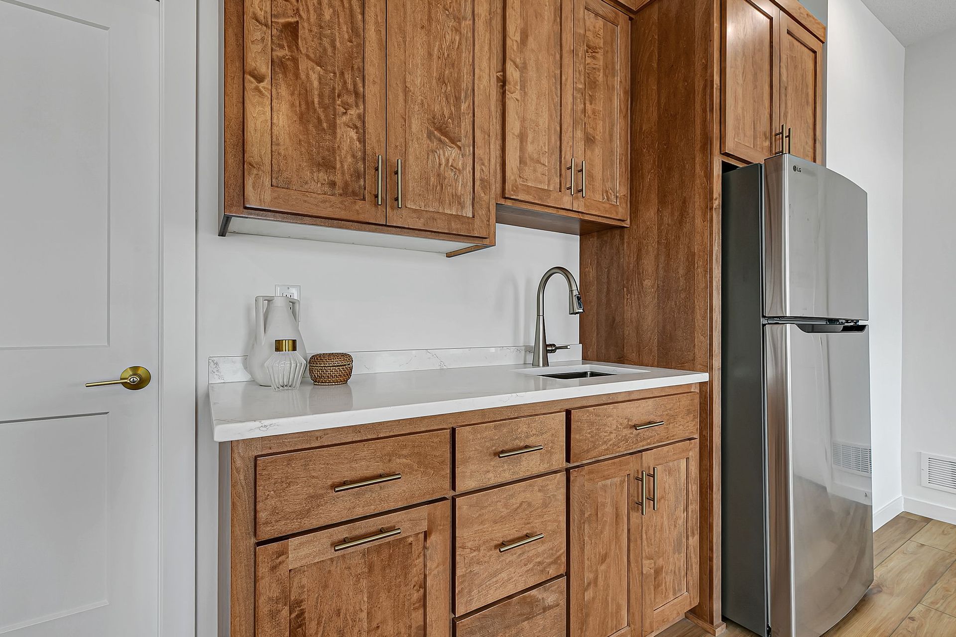 A small kitchenette with wood cabinets, white countertops, a sink, and a stainless steel refrigerator.