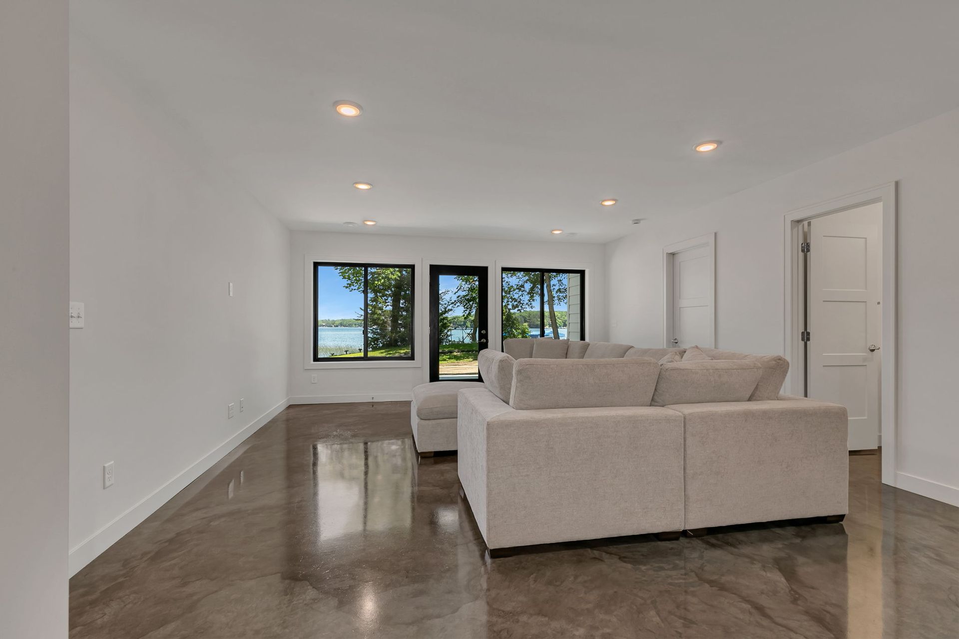 Bright empty living room with white walls, dark floors, beige sofa, and windows at the far end.