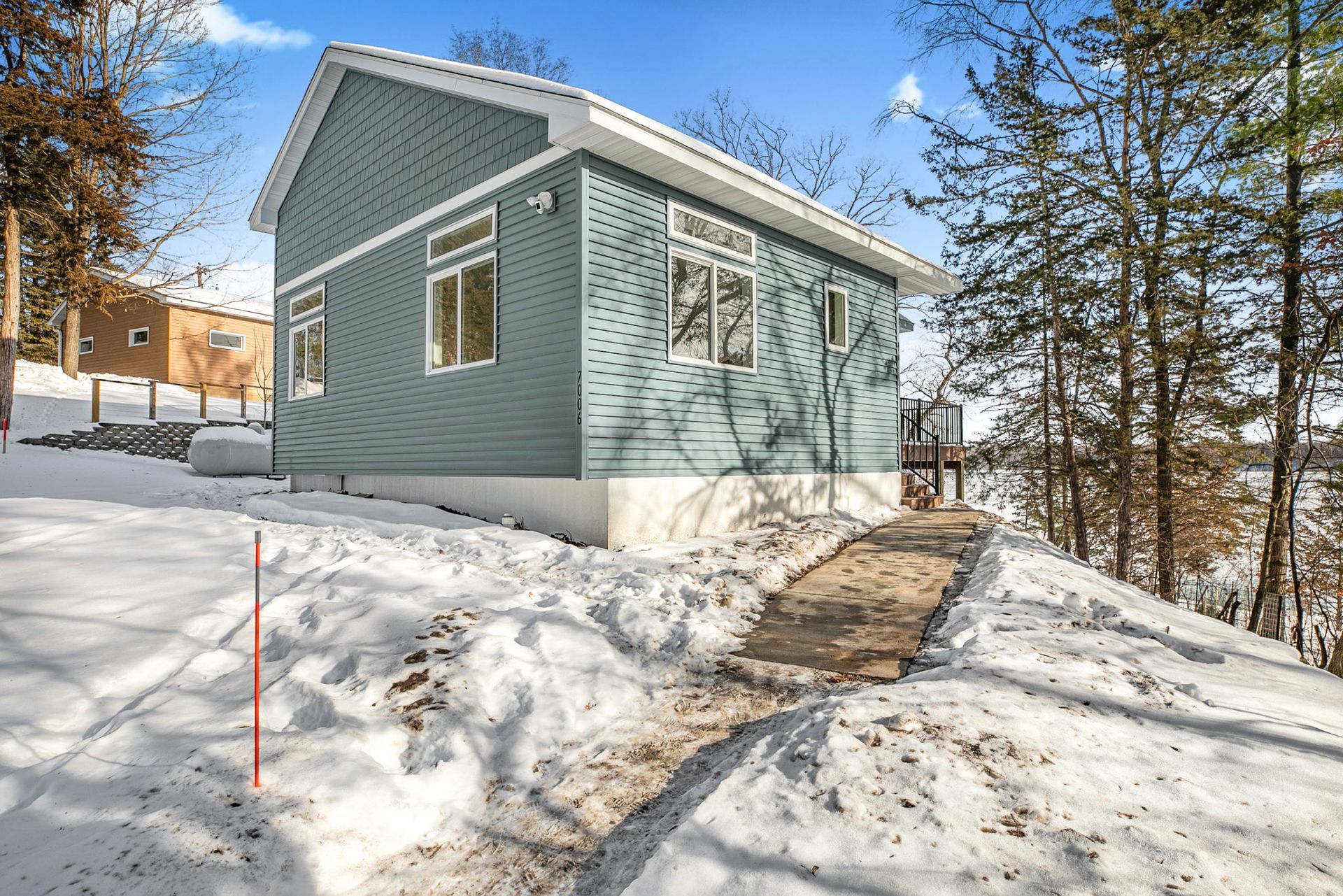 Blue-sided house on snowy hill; a pathway leads to the front. Trees and a lake are in the background.