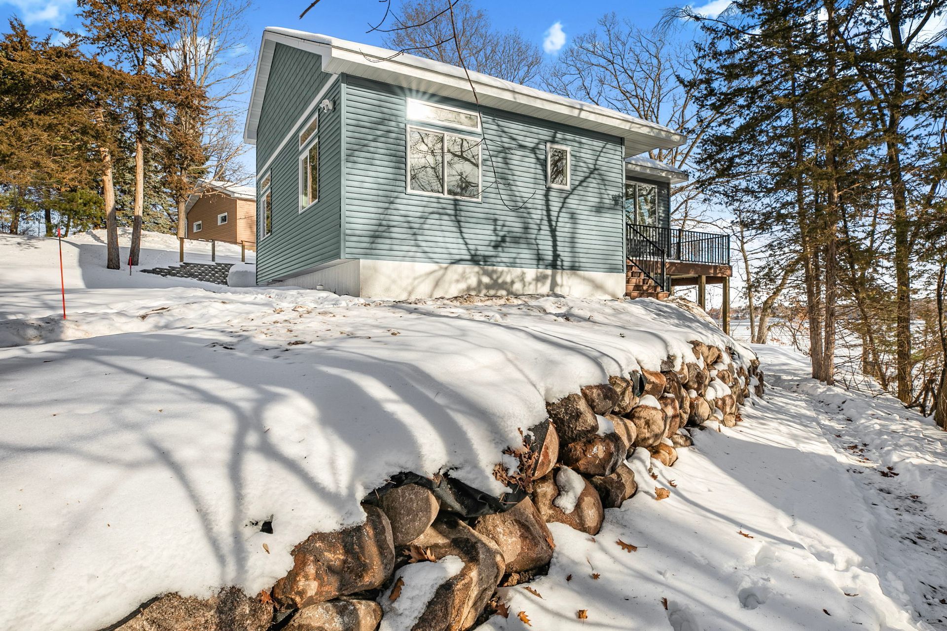 Small blue house on a snowy hill, rock wall in foreground, trees in background.