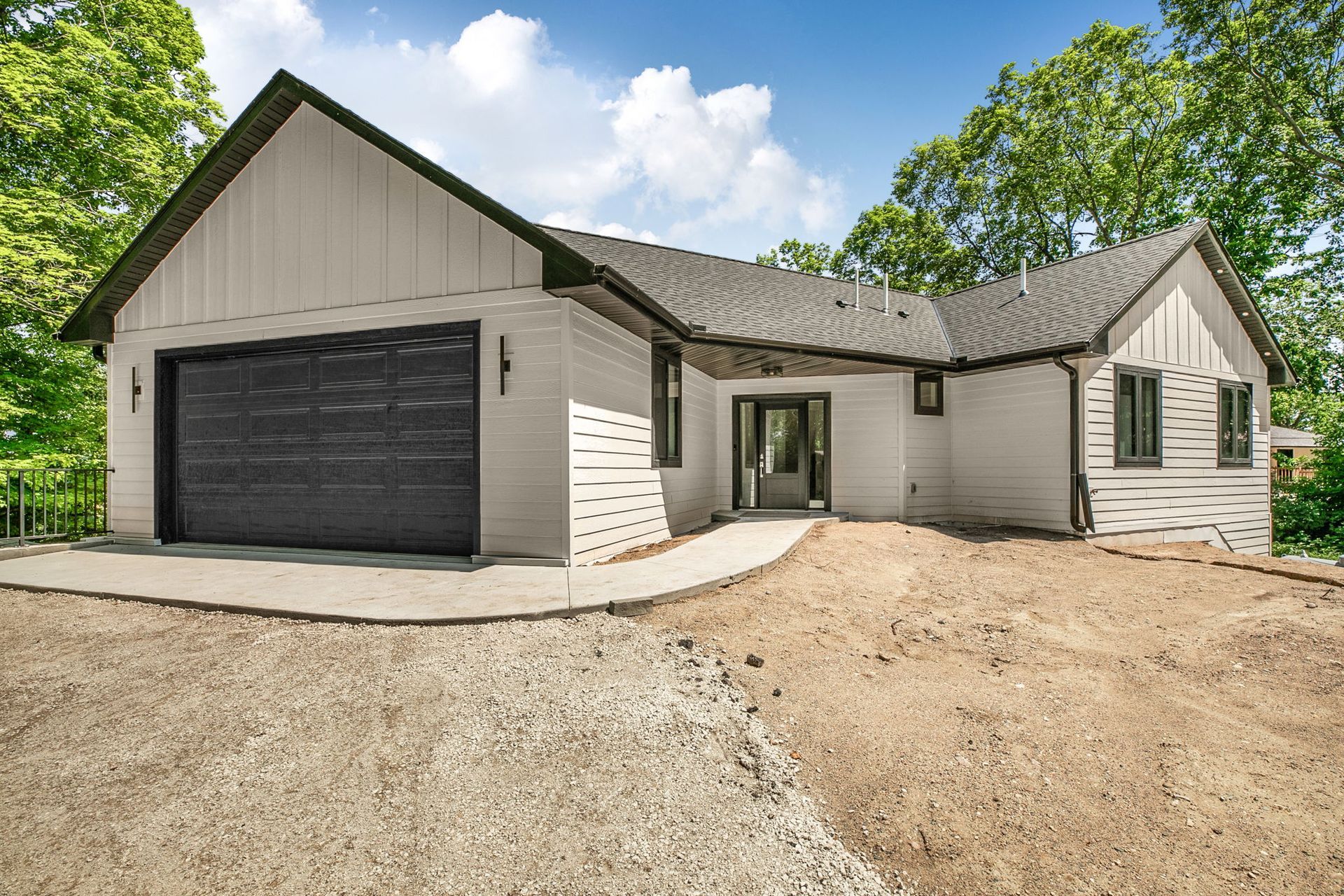 Modern white house with dark garage door, gravel driveway, and trees under a partly cloudy sky