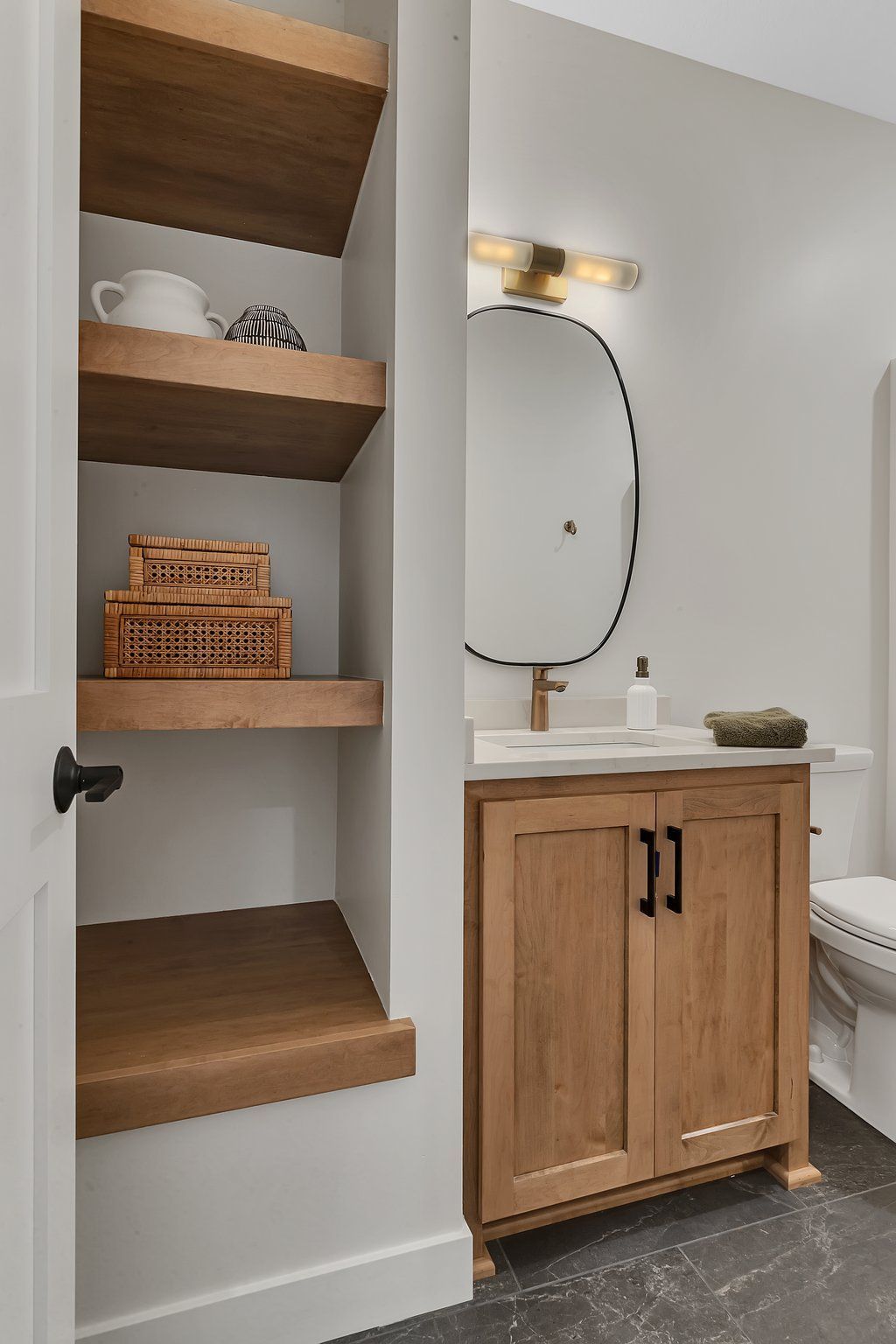 A bathroom with a sink , toilet , mirror and shelves.