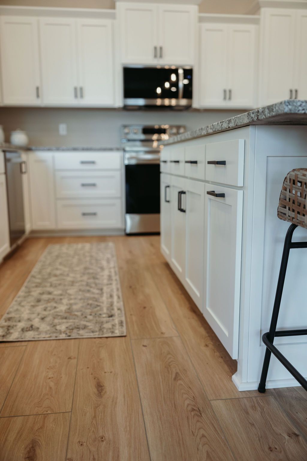 Kitchen with white cabinets, stainless steel appliances, and wooden flooring. A rug and a bar stool are visible.