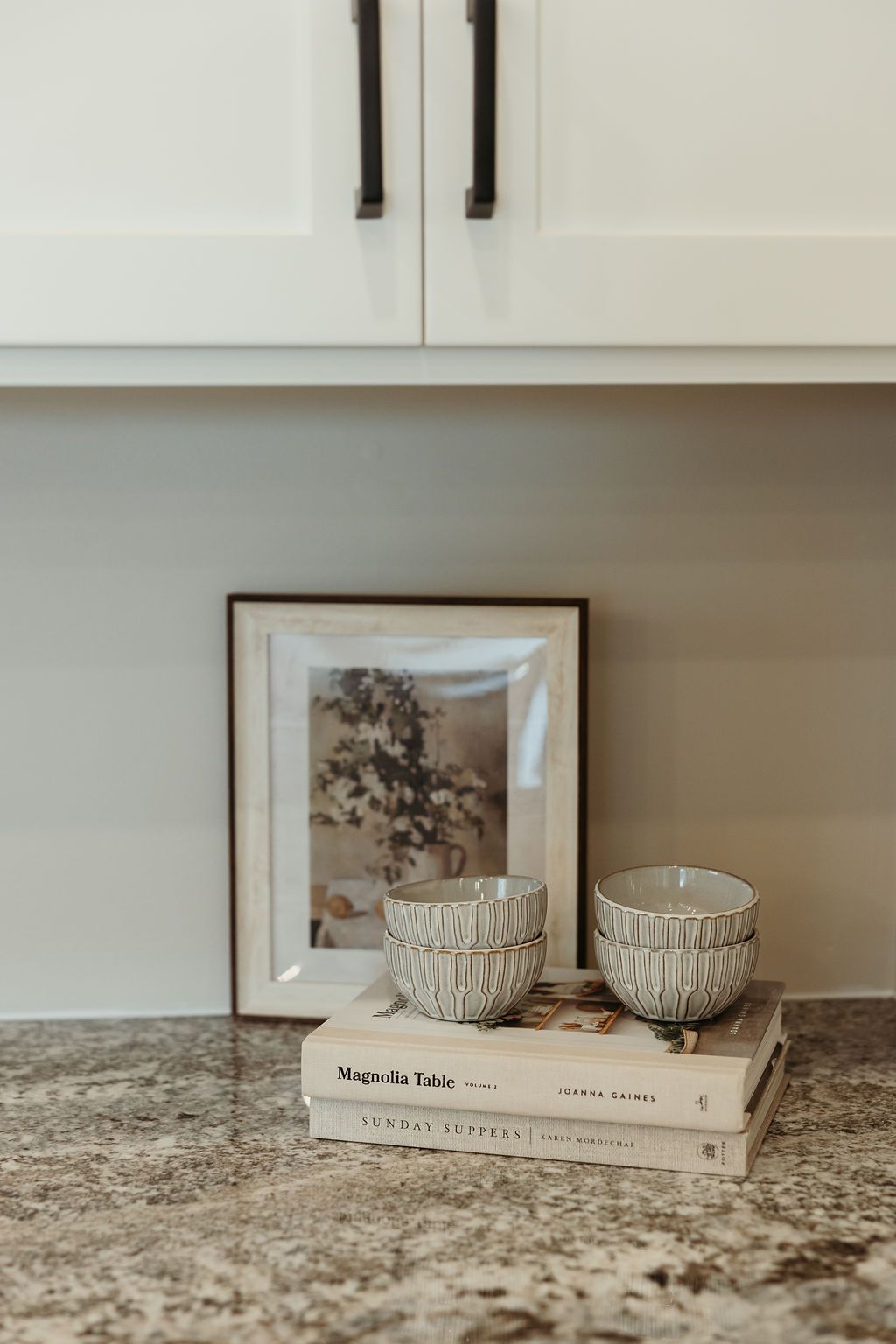 Decorated countertop with framed artwork, stacked books, and decorative bowls under white cabinets with black handles.