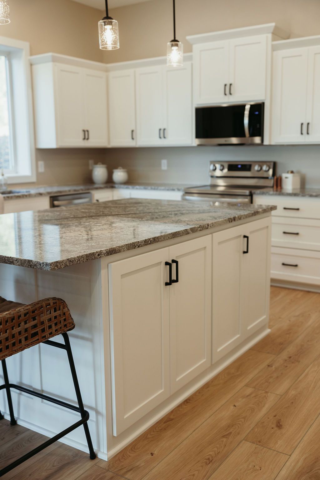 White kitchen with granite countertops, island with black handles, light wood flooring, and pendant lights.