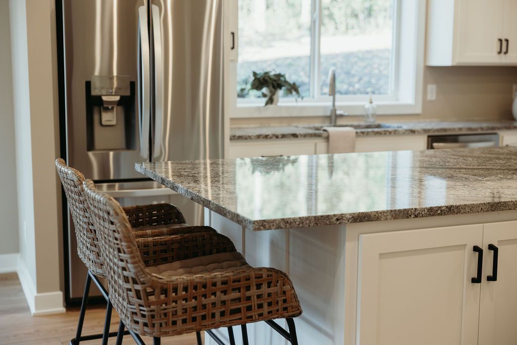 Kitchen island with granite countertop, wicker bar stools, stainless steel refrigerator, and a window overlooking a yard.