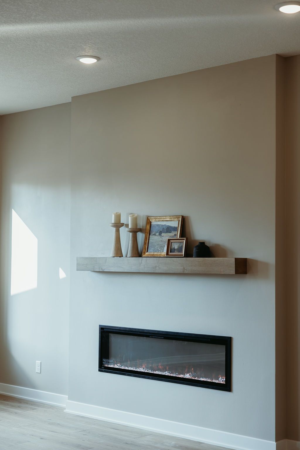 Modern fireplace with floating wooden shelf, candles, and framed art against a light gray wall.