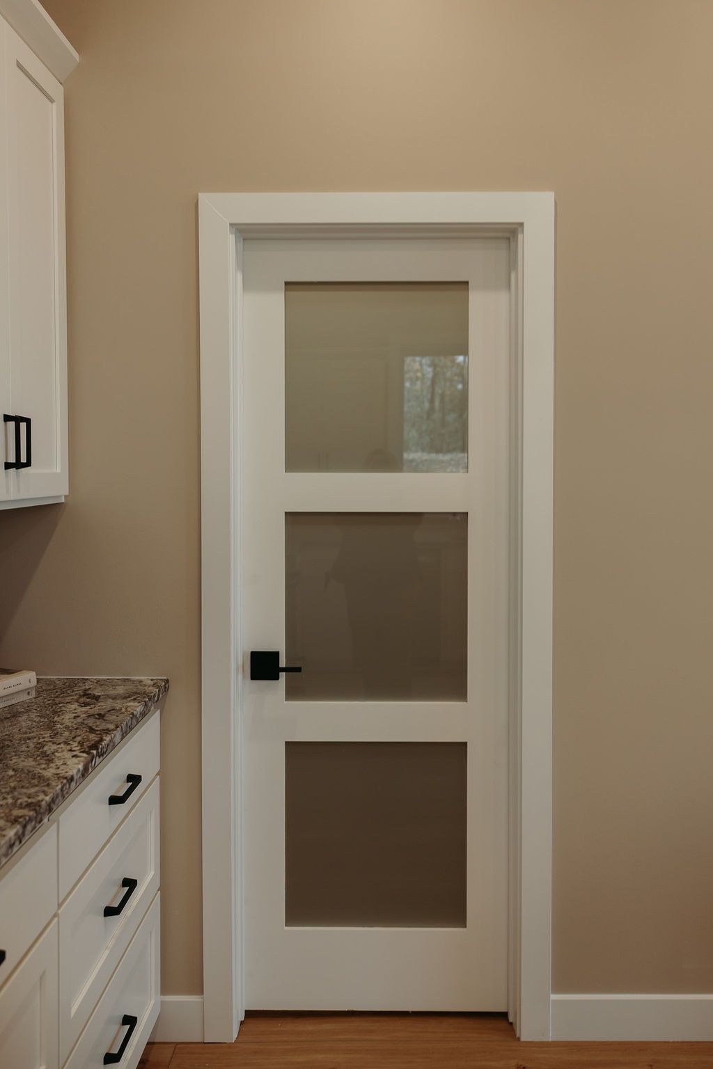 White door with three glass panels, black handle, set in a white frame against a tan wall.