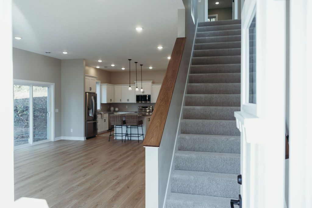 Interior view of a modern home with a staircase, kitchen, and living area. Light wood floors, white walls.