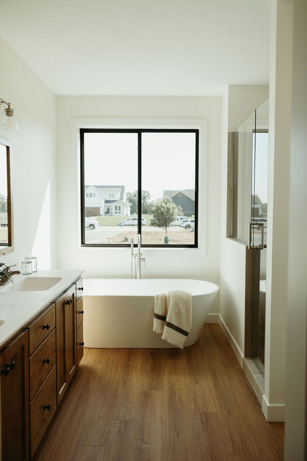 Bathroom with a white bathtub, dark-framed window, wooden floor, and a double vanity with a mirror.