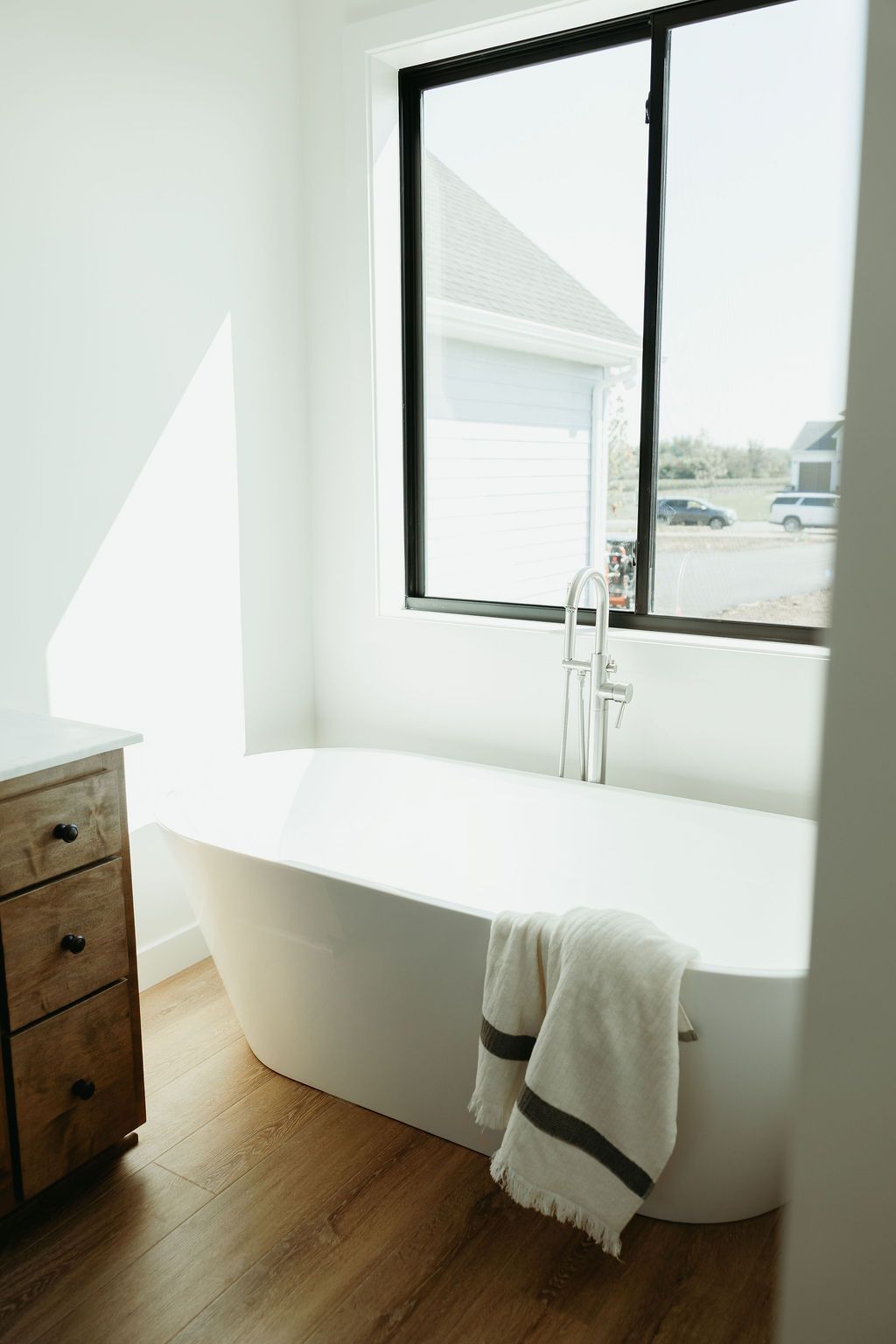 A white bathtub in a modern bathroom near a window, towel draped on side, wooden vanity.