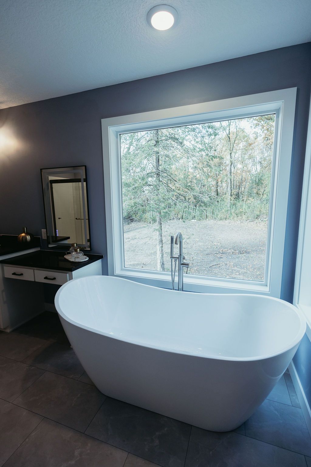 White freestanding bathtub in front of a large window overlooking a wooded area. Bathroom with gray walls.