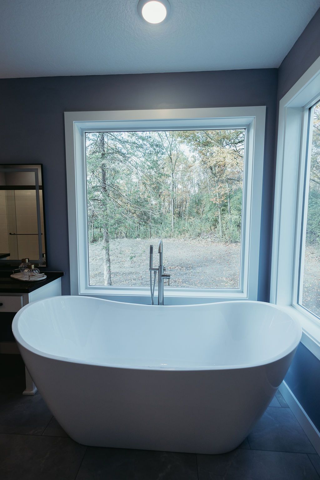 Freestanding white bathtub in a bathroom with a large window overlooking trees.