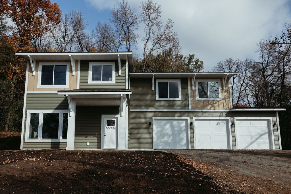 Two-story house with green and tan siding, white garage doors, and a forest backdrop.