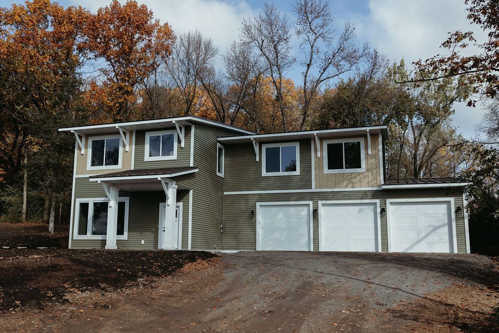Two-story house with three-car garage. Exterior has green and beige siding with white trim. Autumn trees in the background.