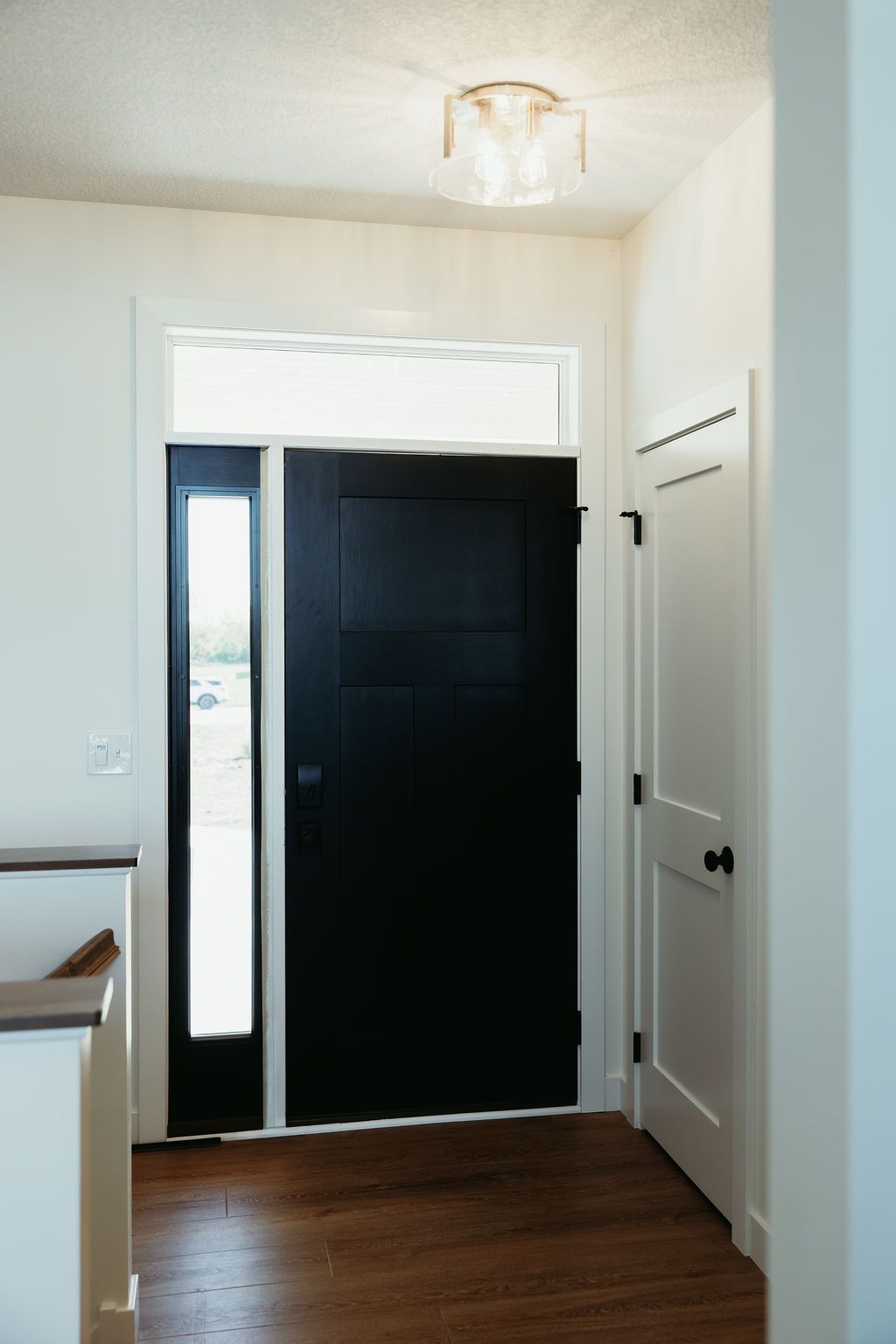 Black front door with sidelight and white trim, wood floor, light fixture, and white walls.