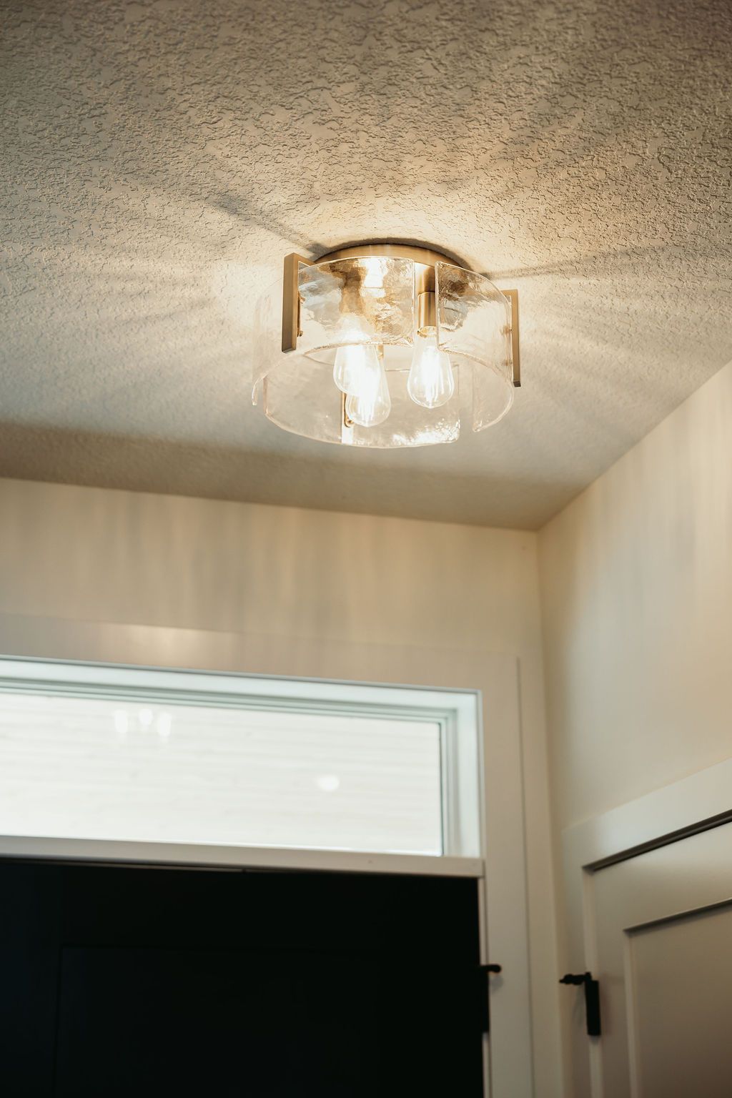 Flush-mount ceiling light fixture with glass shade and gold accents. Below, a window and white walls.