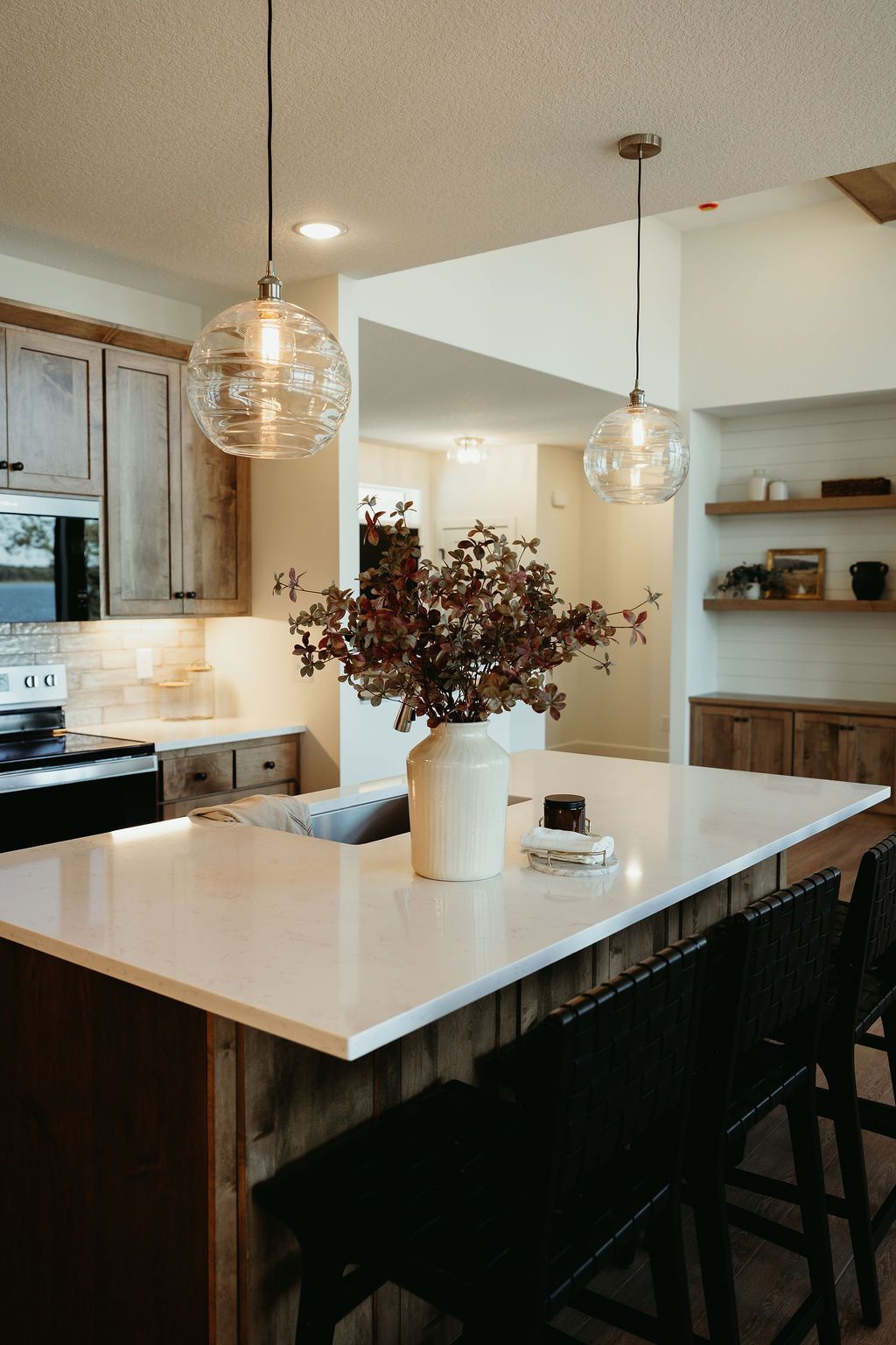 Kitchen with white countertops, wooden cabinets and island, two globe pendant lights, and a vase of flowers.