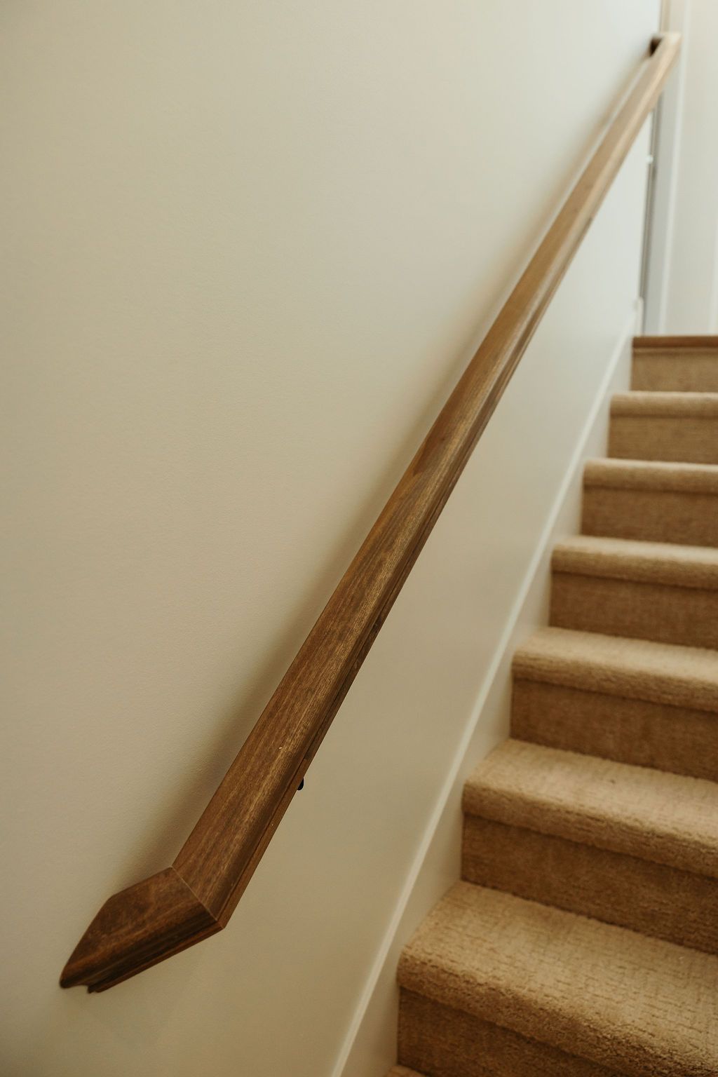 Wooden handrail on a beige wall alongside carpeted stairs.