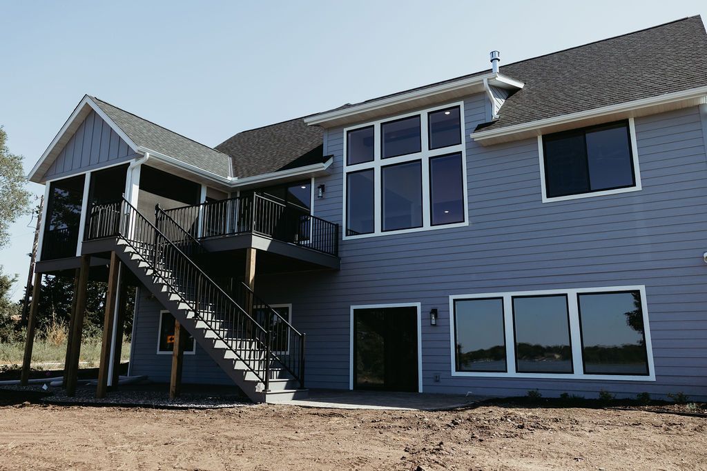 Back of a two-story blue house with large windows, a deck, and stairs leading down to a dirt yard.