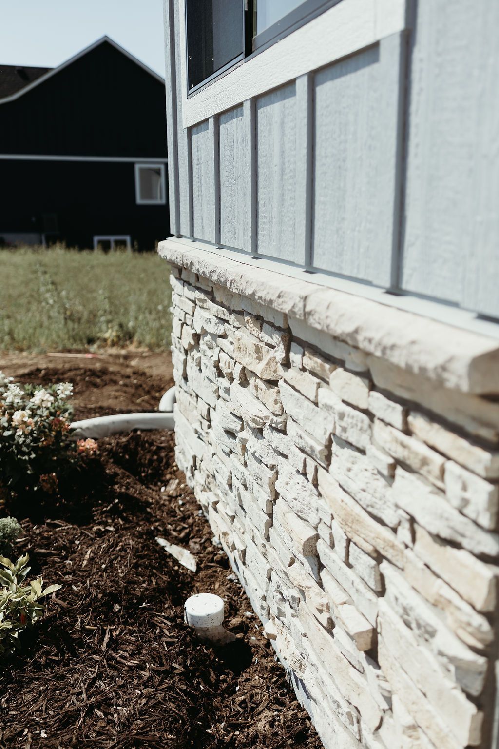 Exterior wall with stone base and light blue siding, with dark mulch and a house in the background.