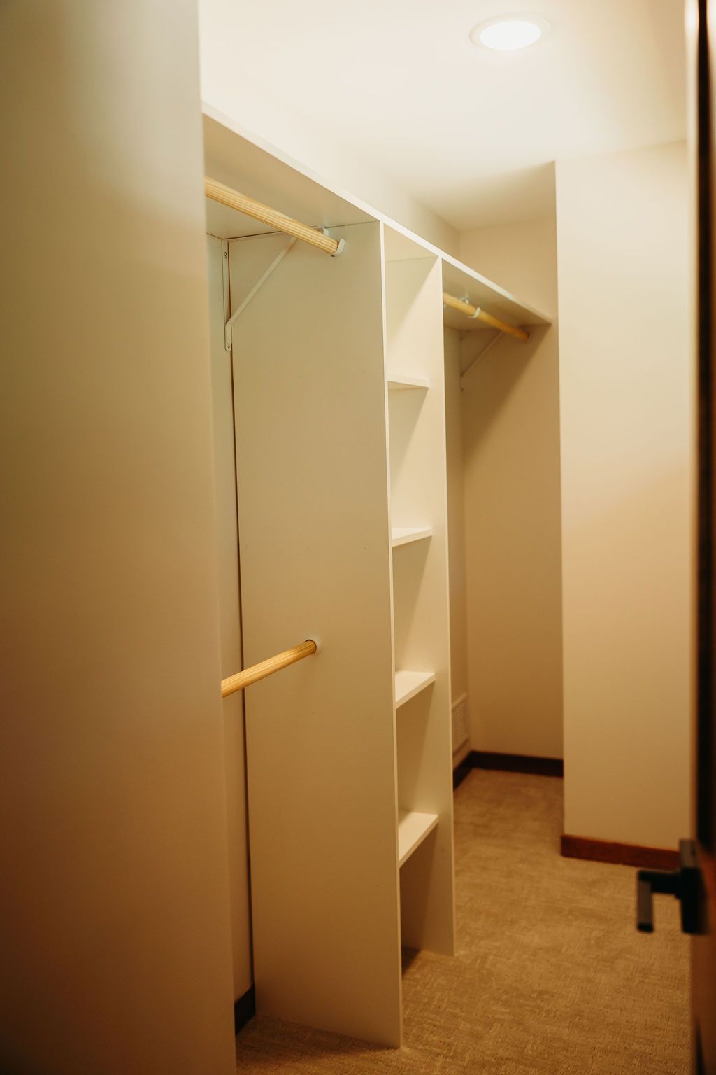 Empty walk-in closet with white shelves and wooden rods, carpeted floor.