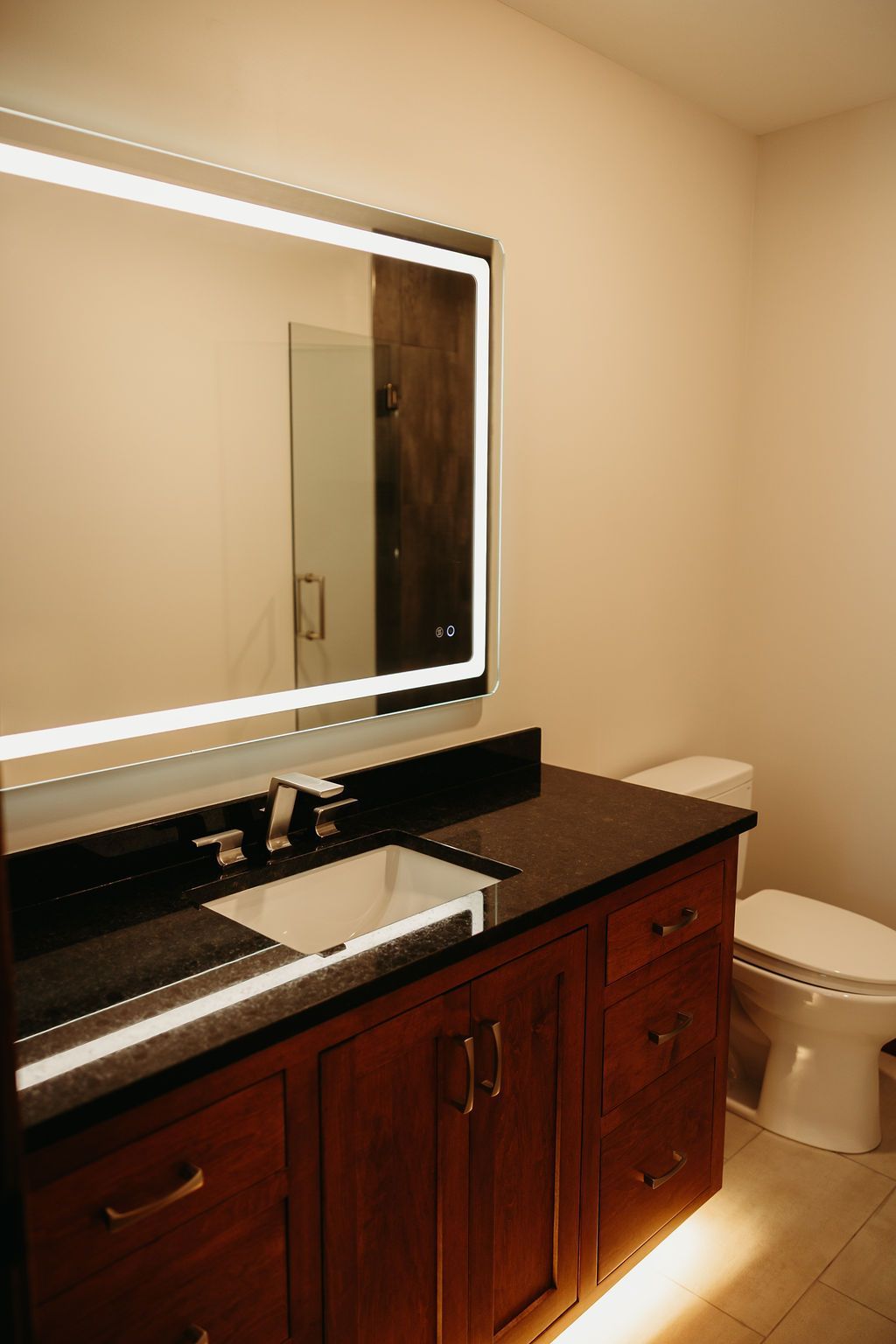 Bathroom with a dark countertop, wooden cabinets, a lit mirror, and a toilet.
